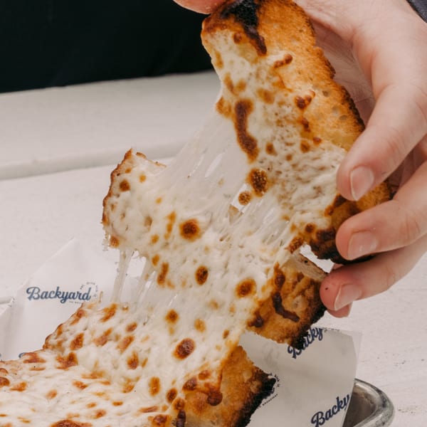 Hands pulling apart cheesy garlic bread, creating long, gooey cheese strings, with "Backyard" branding visible.